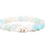 A beaded stretch Fahlo bracelet featuring light blue, white, and pale yellow-green gradient beads, with a small lion charm at the center, set against a plain white background. Available at Be Charmed