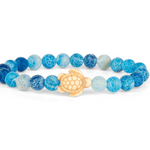 A beaded stretch Fahlo bracelet featuring textured blue and light blue round beads with a natural wood turtle charm in the center, displayed on a clean white background. Available at Be Charmed