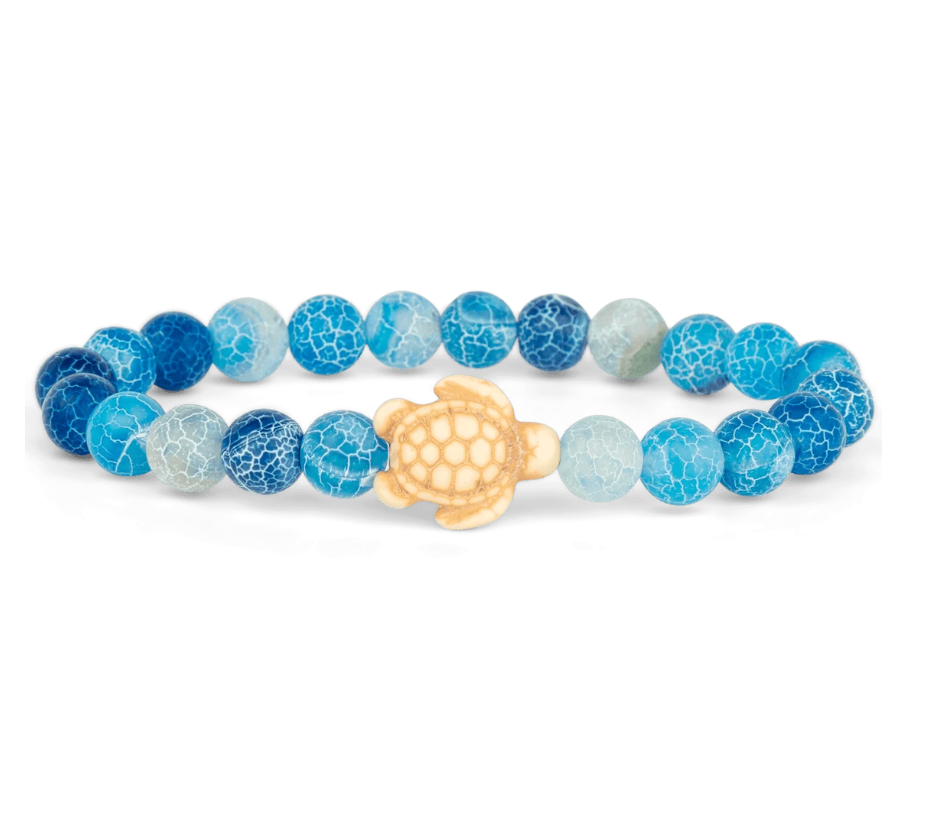 A beaded stretch Fahlo bracelet featuring textured blue and light blue round beads with a natural wood turtle charm in the center, displayed on a clean white background. Available at Be Charmed