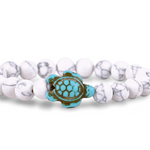 A beaded stretch Fahlo bracelet featuring white and white marbled round beads with a central turquoise and brown turtle charm, shown on a plain white background. Available at Be Charmed