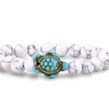 A beaded stretch Fahlo bracelet featuring white and white marbled round beads with a central turquoise and brown turtle charm, shown on a plain white background. Available at Be Charmed