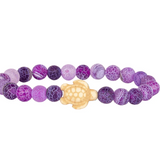A beaded stretch Fahlo bracelet featuring round purple and lavender marbled beads with a textured crackle pattern, accented by a central cream-colored turtle charm, shown on a plain white background. Available at Be Charmed