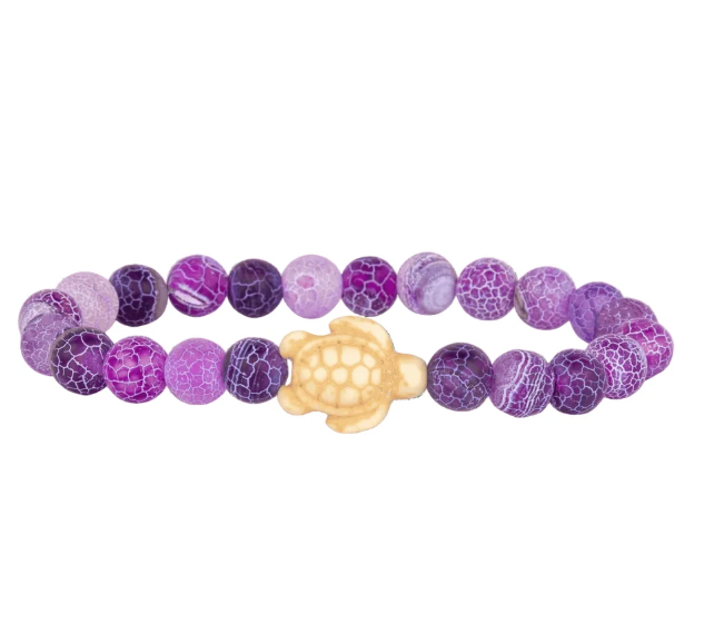 A beaded stretch Fahlo bracelet featuring round purple and lavender marbled beads with a textured crackle pattern, accented by a central cream-colored turtle charm, shown on a plain white background. Available at Be Charmed