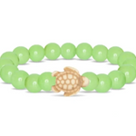 A beaded stretch Fahlo bracelet featuring a sage glow round beads and a central carved turtle charm in a natural tan wood tone, shown on a plain white background. Available at Be Charmed
