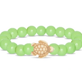 A beaded stretch Fahlo bracelet featuring a sage glow round beads and a central carved turtle charm in a natural tan wood tone, shown on a plain white background. Available at Be Charmed