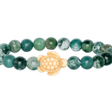 A beaded stretch Fahlo bracelet featuring round seagrass green marbled beads with a central cream-colored turtle charm, shown on a plain white background. Available at Be Charmed