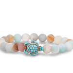 A beaded stretch Fahlo bracelet featuring a central turquoise and bronze sea turtle charm, surrounded by smooth round beads in pastel shades of light blue, cream, peach, and gray on a white background. Available at Be Charmed