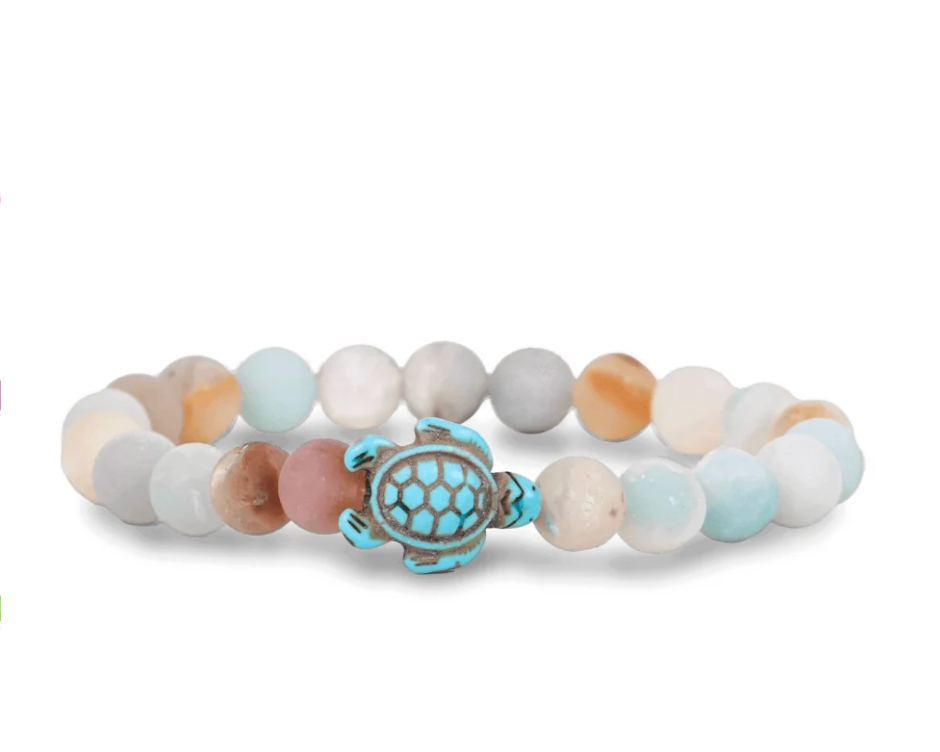 A beaded stretch Fahlo bracelet featuring a central turquoise and bronze sea turtle charm, surrounded by smooth round beads in pastel shades of light blue, cream, peach, and gray on a white background. Available at Be Charmed