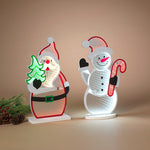 Two illuminated Christmas LED light sculptures shaped like Santa Claus and a snowman, displayed on a beige background with pine branches and pinecones at the side. Availabe at Be Charmed