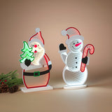 Two illuminated Christmas LED light sculptures shaped like Santa Claus and a snowman, displayed on a beige background with pine branches and pinecones at the side. Availabe at Be Charmed