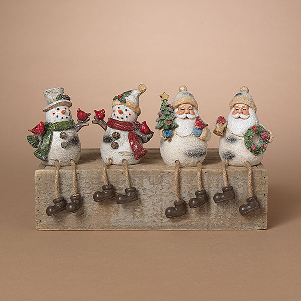 Four Christmas-themed resin figurines sitting on a weathered wooden block against a solid beige background. The set includes two snowmen and two Santa Claus figures, each with painted white bodies, colorful scarves and hats, and dark brown boots. Availabe at Be Charmed