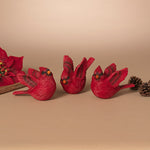 Three red cardinal bird figurines with textured feathers and small orange beaks, arranged on a beige surface with a floral prop and pinecones at the sides. Available at Be Charmed
