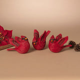 Three red cardinal bird figurines with textured feathers and small orange beaks, arranged on a beige surface with a floral prop and pinecones at the sides. Available at Be Charmed