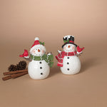 Two decorative snowman figurines on a warm beige background, one with a red Santa hat and green scarf and one with a black top hat and red scarf, styled with cinnamon sticks and pinecones. Available at Be Charmed