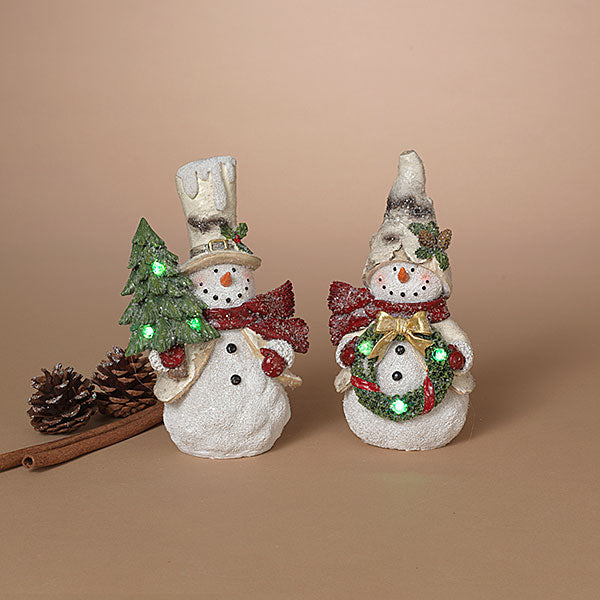 Two decorative snowman figurines with festive holiday accessories, including a small lit Christmas tree, pinecones, and a cinnamon stick, set against a solid beige background. Available at Be Charmed