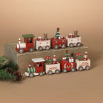 A decorative Christmas train set with red, white, and green painted wooden cars, displayed on a rustic wooden block with pine branches and pinecones against a warm beige background. Available at Be Charmed
