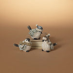 Three small bird figurines dressed in winter clothing, arranged on stacked books against a warm beige gradient background. Available at Be Charmed