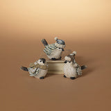Three small bird figurines dressed in winter clothing, arranged on stacked books against a warm beige gradient background. Available at Be Charmed