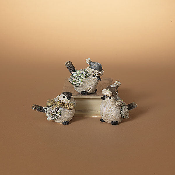 Three small bird figurines dressed in winter clothing, arranged on stacked books against a warm beige gradient background. Available at Be Charmed