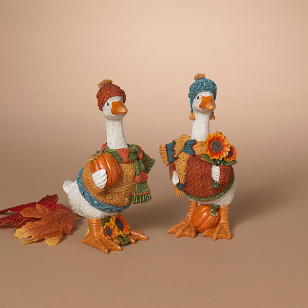 Two decorative autumn goose figurines dressed in knit sweaters, hats, and holding pumpkins and sunflowers, displayed on a warm beige background with a few scattered fall leaves. Available at Be Charmed