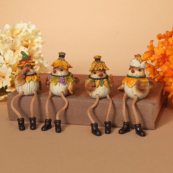 Four small decorative bird figurines dressed in autumn clothing, seated on a wooden block against a warm beige background with floral props on both sides. Available at Be Charmed