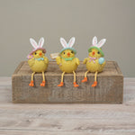 Three Easter chick figurines wearing bunny hats sit on a rustic wooden block against a neutral gray background. Available at Be Charmed