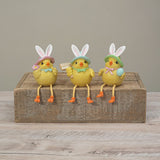 Three Easter chick figurines wearing bunny hats sit on a rustic wooden block against a neutral gray background. Available at Be Charmed