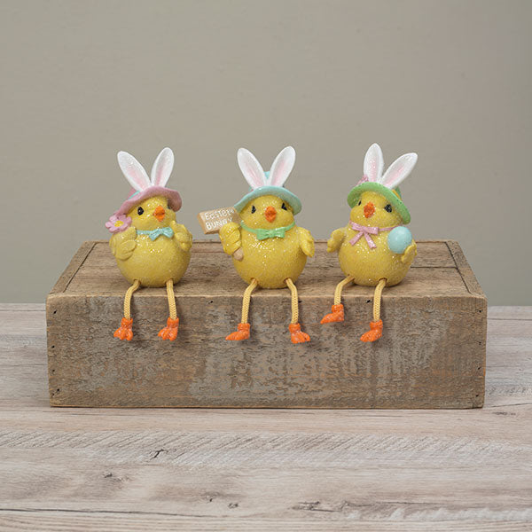 Three Easter chick figurines wearing bunny hats sit on a rustic wooden block against a neutral gray background. Available at Be Charmed