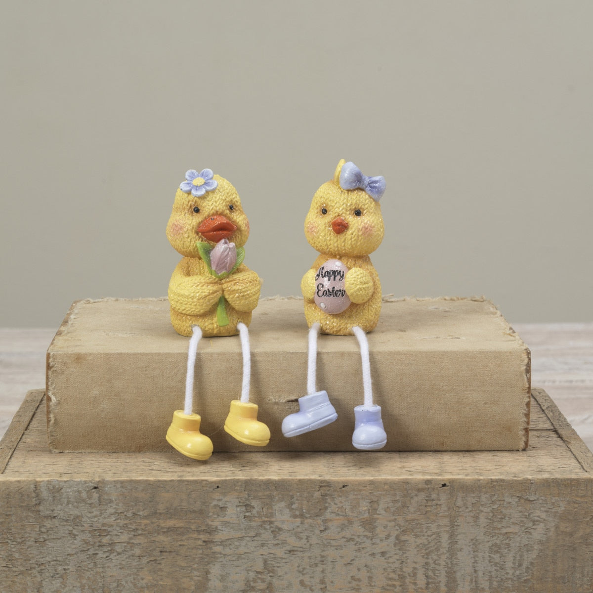 Two small Gerson yellow chick figurines sitting on a rustic wooden block against a neutral beige background. One chick holds a purple flower and wears yellow boots, while the other wears light blue boots and holds a sign that says 'Happy Easter'. Available at Be Charmed