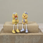 Two small Gerson yellow chick figurines sitting on a rustic wooden block against a neutral beige background. One chick holds a purple flower and wears yellow boots, while the other wears light blue boots and holds a sign that says 'Happy Easter'. Available at Be Charmed