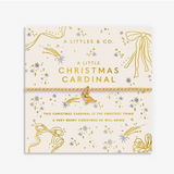A Little Christmas Cardinal bracelet by A Little's & Co. displayed on its cream-colored illustrated card packaging, featuring gold and silver glittery details and a small gold bird charm. Available at Be Charmed