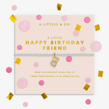A silver-toned bracelet with a gold heart charm and rhinestone trim, presented on a pale pink card that reads 'A LITTLE HAPPY BIRTHDAY FRIEND.' The card is surrounded by pink and gold confetti on a white background. Available at Be Charmed