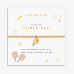 A gold pickleball bracelet by A Littles & Co. on a white gift card with gold and pink text and illustrations, featuring a small pickleball paddle charm. Available at Be Charmed