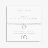 A silver chain bracelet with a small ring charm and tiny diamond-like accents, displayed on a white card that reads 'A LITTLE YOU'RE ENGAGED!' with wedding ring graphics. Brand 'A LITTLES & CO. Available at Be Charmed
