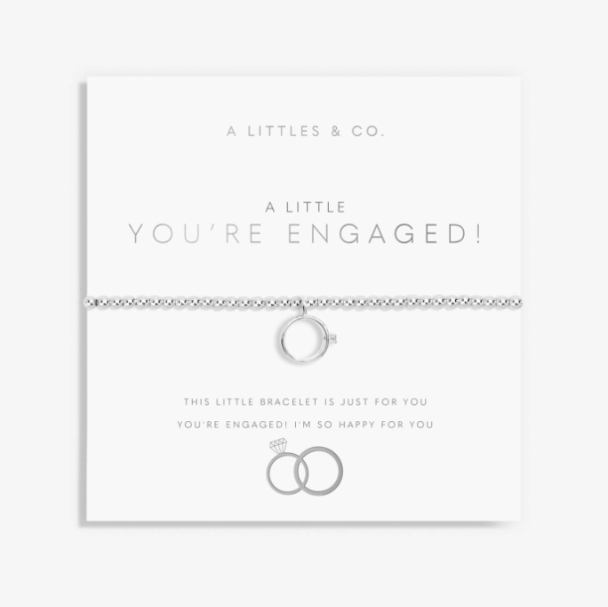 A silver chain bracelet with a small ring charm and tiny diamond-like accents, displayed on a white card that reads 'A LITTLE YOU'RE ENGAGED!' with wedding ring graphics. Brand 'A LITTLES & CO. Available at Be Charmed