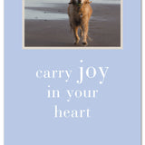 A greeting card featuring an image of a dog on the beach with the text 'carry joy in your heart' printed on it. Available at Be Charmed
