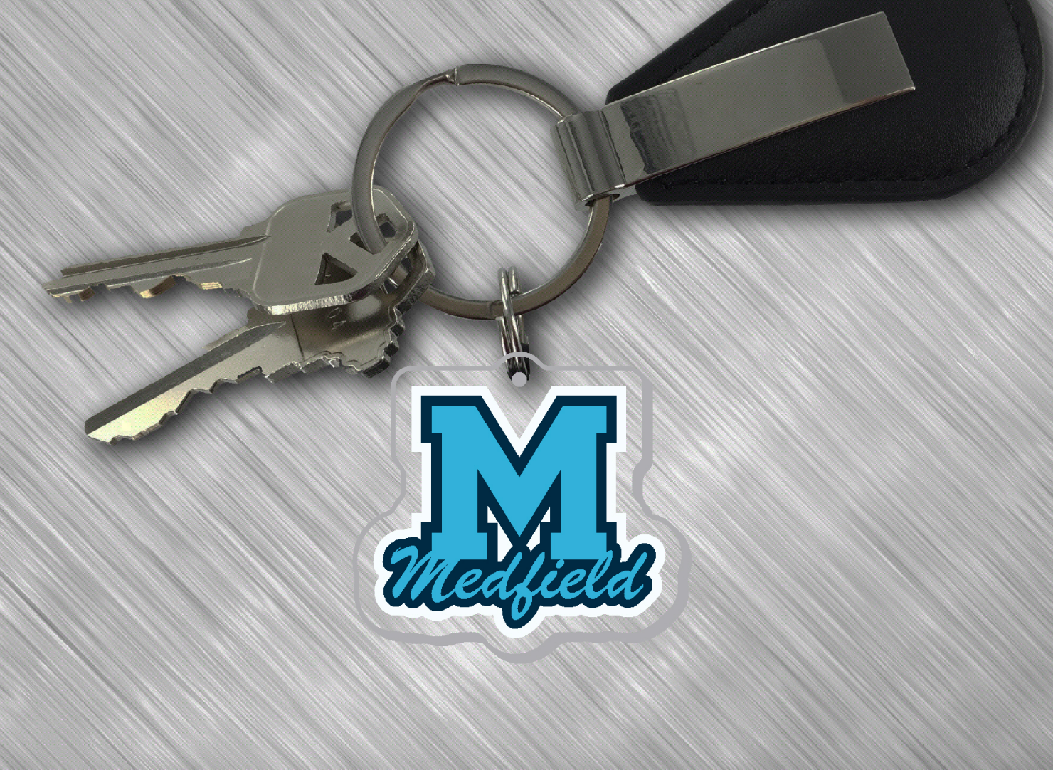 A clear acrylic keychain with a metal ring, featuring a blue 'M' logo that reads 'MEDFIELD', attached to a black keychain clip. Available at Be Charmed