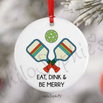 A white ceramic Christmas ornament with a red hanging ribbon, featuring a crossed-paddle tennis design with a green patterned ball and the text 'EAT, DINK & BE MERRY'. Available at Be Charmed