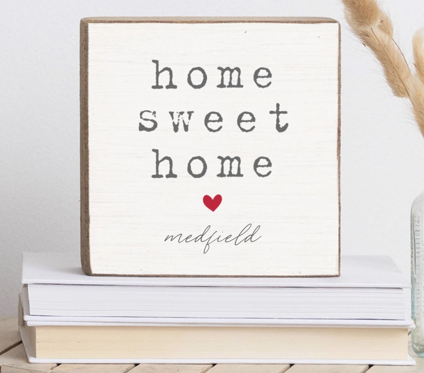 A Rustic Marlin square decorative wooden block with the phrase 'home sweet home' in dark gray lettering, a small red heart, and the name 'medfield' in script at the bottom. The sign is stacked on books with dried grass stems in the background. Available at Be Charmed