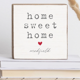 A Rustic Marlin square decorative wooden block with the phrase 'home sweet home' in dark gray lettering, a small red heart, and the name 'medfield' in script at the bottom. The sign is stacked on books with dried grass stems in the background. Available at Be Charmed