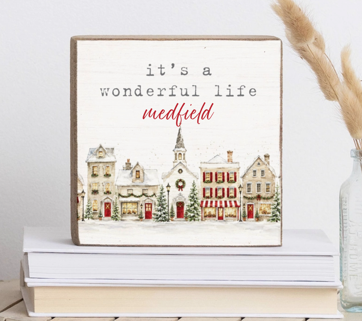 A square decorative block with a rustic wood border and a painted winter village scene. The sign reads “it’s a wonderful life medfield” in black and red lettering.  Available at Be Charmed