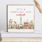 A square decorative block with a rustic wood border and a painted winter village scene. The sign reads “it’s a wonderful life medfield” in black and red lettering.  Available at Be Charmed