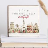 A square decorative block with a rustic wood border and a painted winter village scene. The sign reads “it’s a wonderful life medfield” in black and red lettering.  Available at Be Charmed