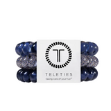 A set of four Teleties hair elastic bands wrapped together with a white paper band. The bands are navy blue with some featuring a textured pattern of navy. Available at Be Charmed