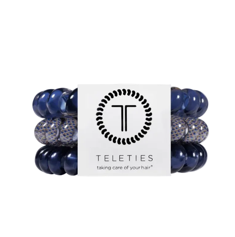 A set of four Teleties hair elastic bands wrapped together with a white paper band. The bands are navy blue with some featuring a textured pattern of navy. Available at Be Charmed