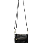 A Joy Susan Lia black pebbled crossbody bag with a long shoulder strap, hanging from a wooden peg against a solid white background. Available at Be Charmed