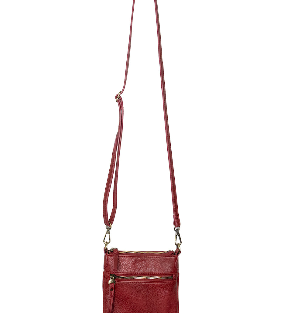 A Joy Susan Lia chilli crossbody bag with a long adjustable strap, hanging from a wooden peg against a plain white background. Available at Be Charmed