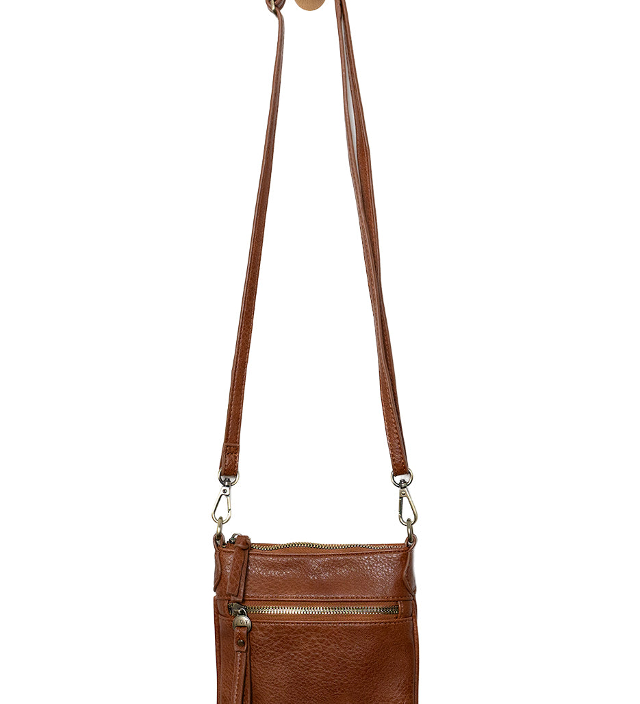 A Joy Susan cognac Lia crossbody bag with a long adjustable strap, hanging from a wooden peg on a white background. Available at Be Charmed
