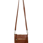 A Joy Susan cognac Lia crossbody bag with a long adjustable strap, hanging from a wooden peg on a white background. Available at Be Charmed