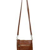 A Joy Susan cognac Lia crossbody bag with a long adjustable strap, hanging from a wooden peg on a white background. Available at Be Charmed